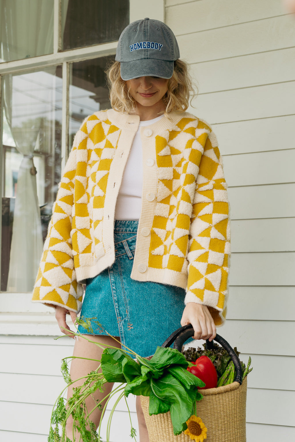 Woman wearing a yellow and white patterned jacket and blue denim skirt, holding a basket of groceries.