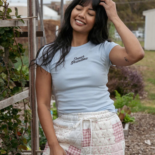 Woman wearing a light blue t-shirt and patterned pants standing in an outdoor garden setting.