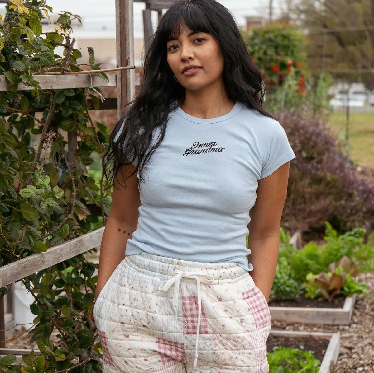 Woman wearing a light blue t-shirt with text and patterned pants standing in a garden.