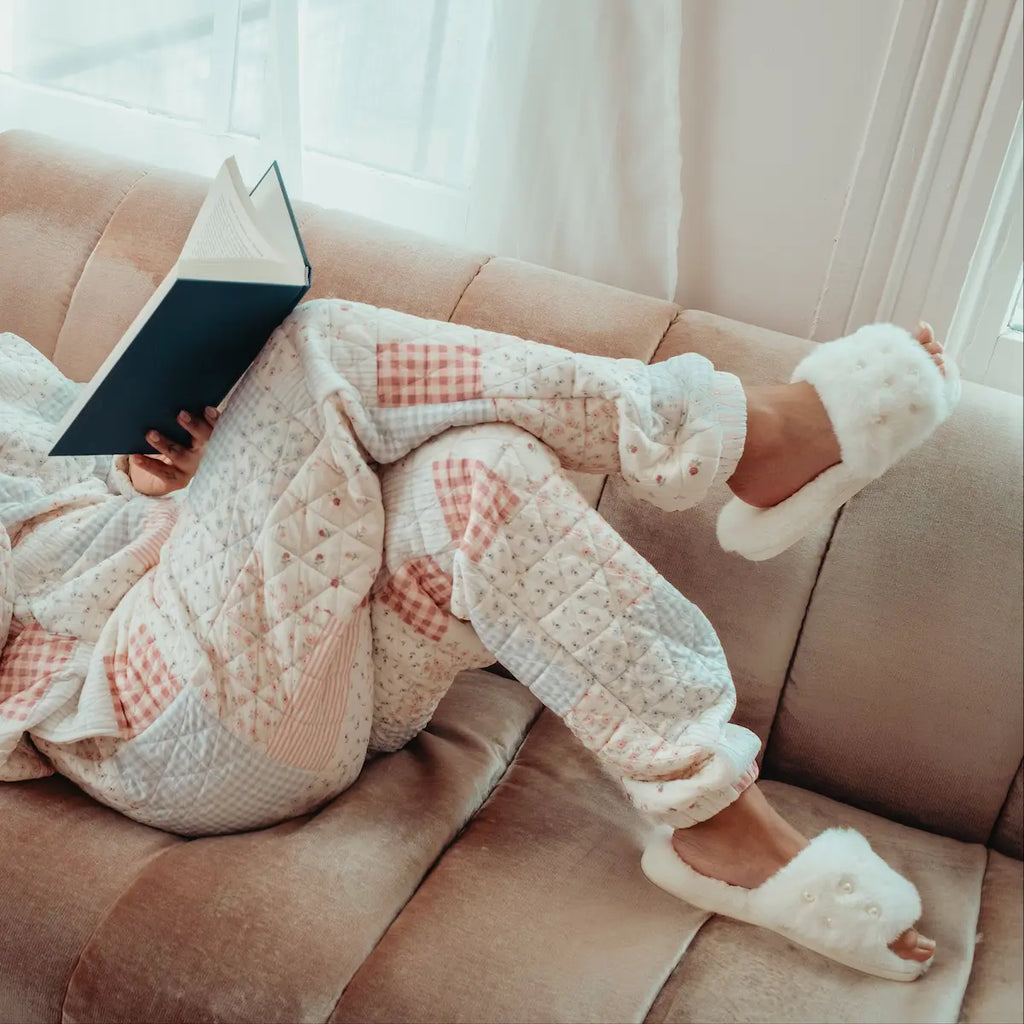 pajamas reading a book on a couch in a cozy living room with white open toe slippers with pearl details