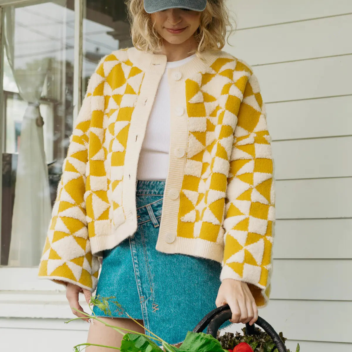 Woman wearing a yellow and white patterned jacket and blue denim skirt, holding a basket of groceries.