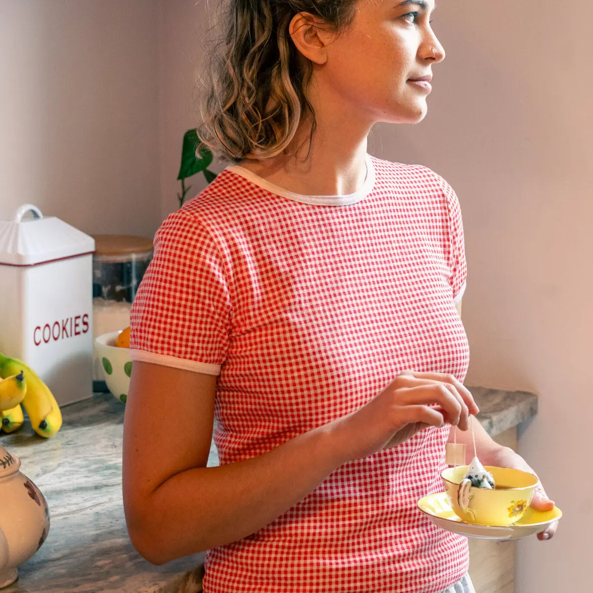 Woman in a kitchen wearing a red gingham tee holding a teacup. 