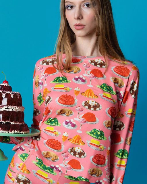 Person wearing a pink long sleeve with dessert patterns, holding a cake against a blue background
