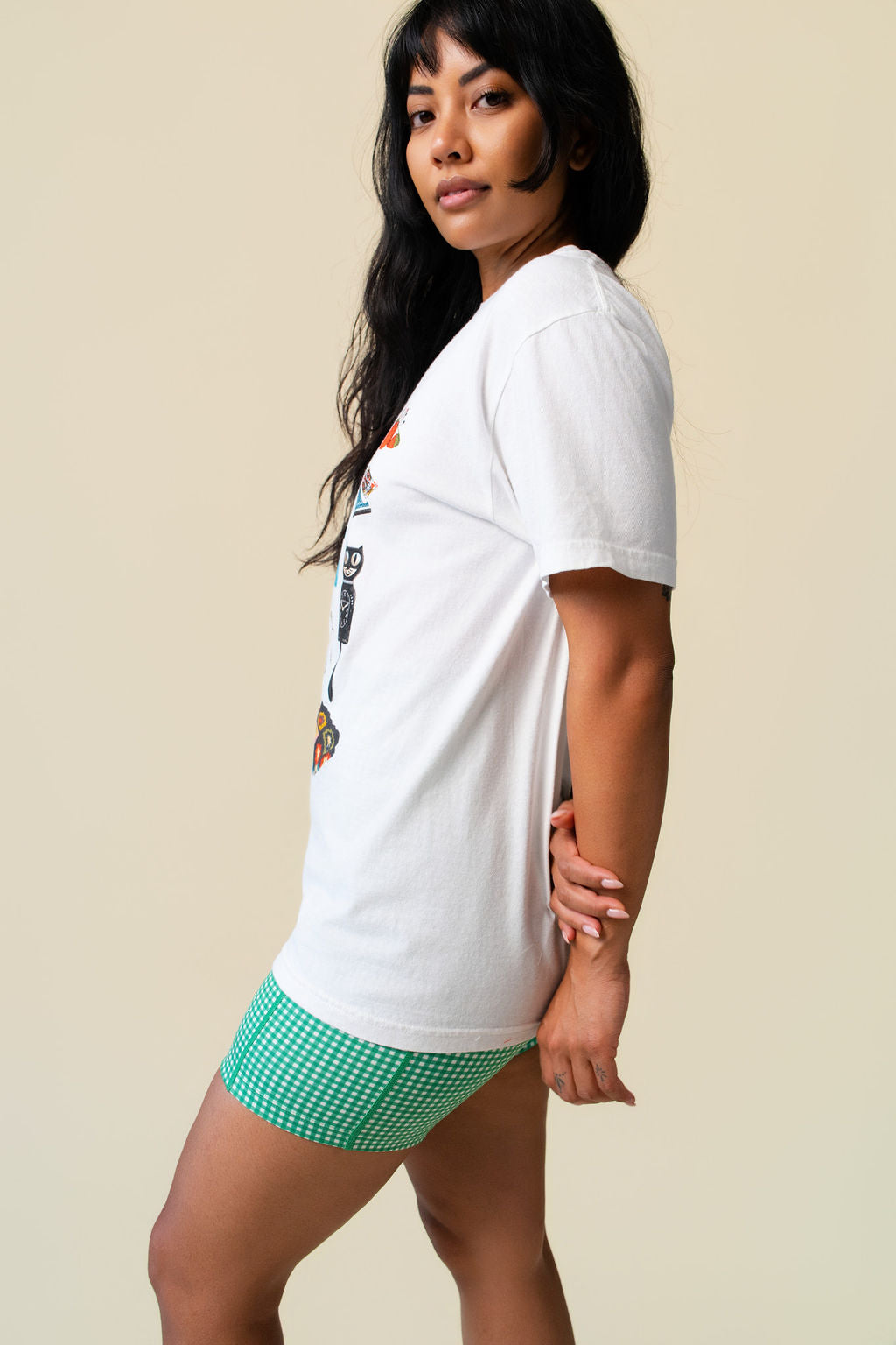 Side of person wearing a white t-shirt with colorful graphics of grandma's items with green gingham biker shorts on a beige background.