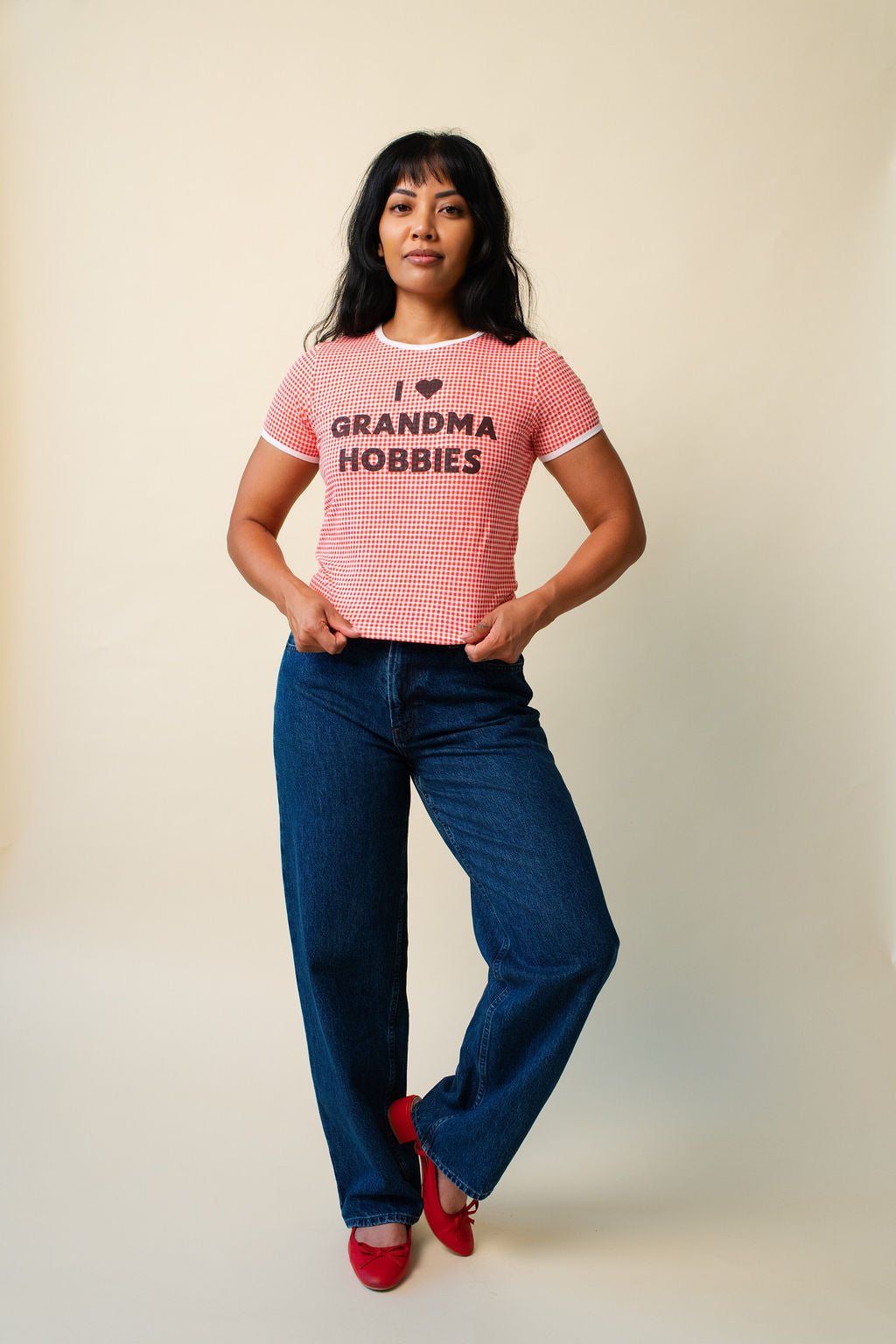 Person wearing a red and white checkered shirt with 'I ❤️ Grandma Hobbies' text with jeans and red shoes on a beige background.