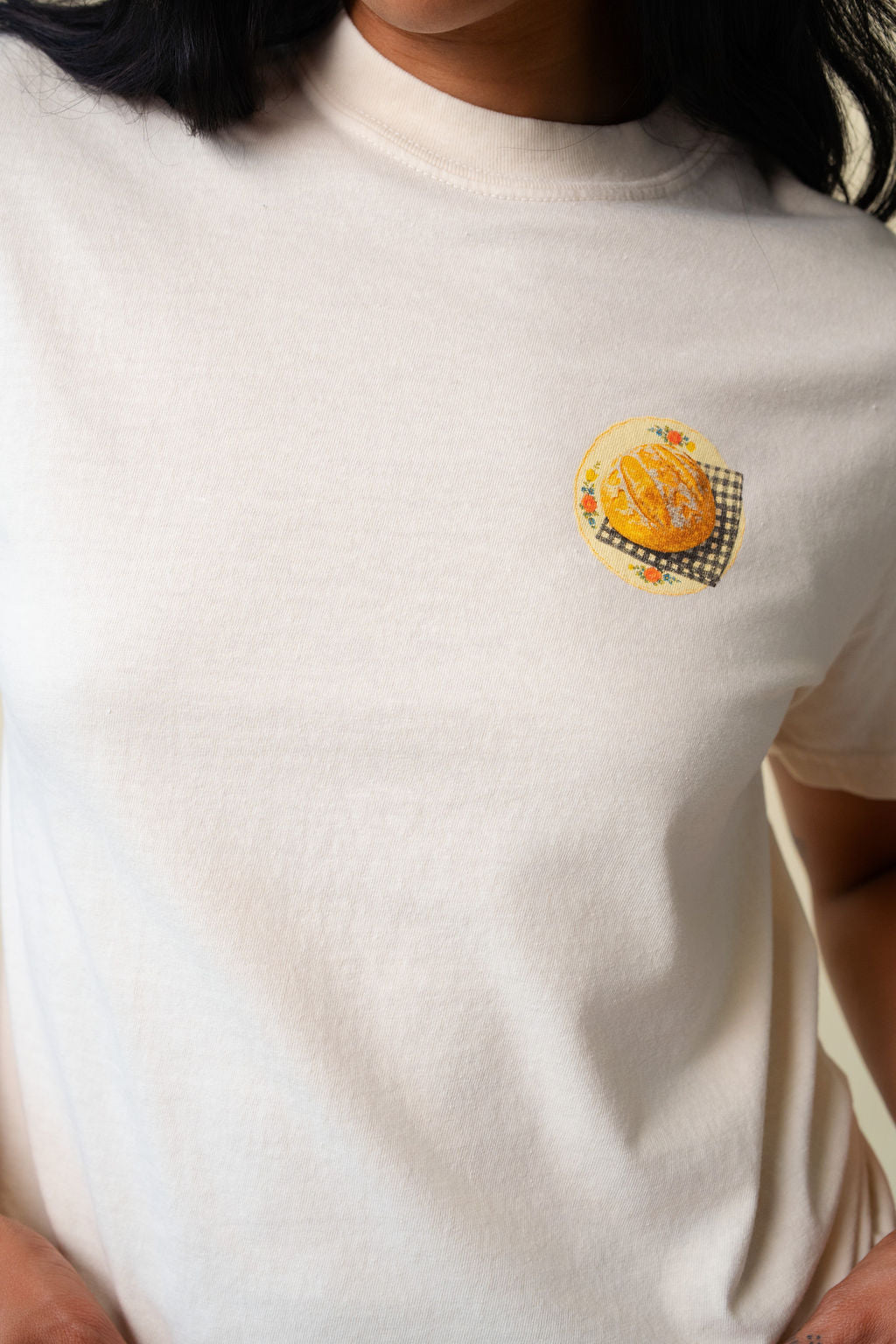 Close up of a person wearing a beige t-shirt with a small graphic of sour dough bread.