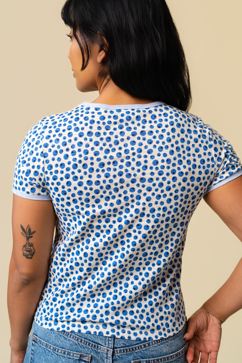 Back of person wearing a blueberry printed short sleeve paired with blue jeans standing on a beige background.