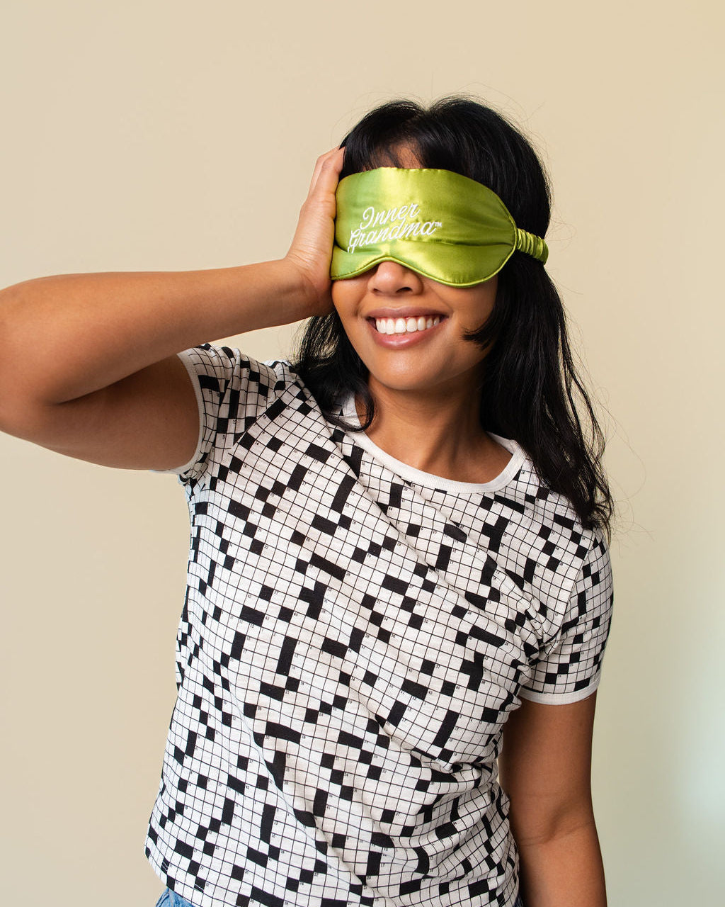 A woman wearing a black and white cross-word pattern t-shirt and blue jeans, smiling while wearing a lime green satin sleep mask with the text 'Inner Grandma' on it, against a plain beige background.