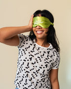 A woman wearing a black and white cross-word pattern t-shirt and blue jeans, smiling while wearing a lime green satin sleep mask with the text 'Inner Grandma' on it, against a plain beige background.