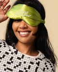 A woman wearing a shiny olive green satin sleep mask with 'Inner Grandma' embroidered in white script, paired with a black and white patterned top against a solid beige background.