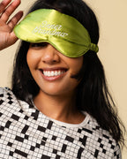 A woman wearing a shiny olive green satin sleep mask with 'Inner Grandma' embroidered in white script, paired with a black and white patterned top against a solid beige background.