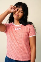 Person wearing a red and white gingham printed tee with a red and white "Tired" patch on the front, rubbing their eye.