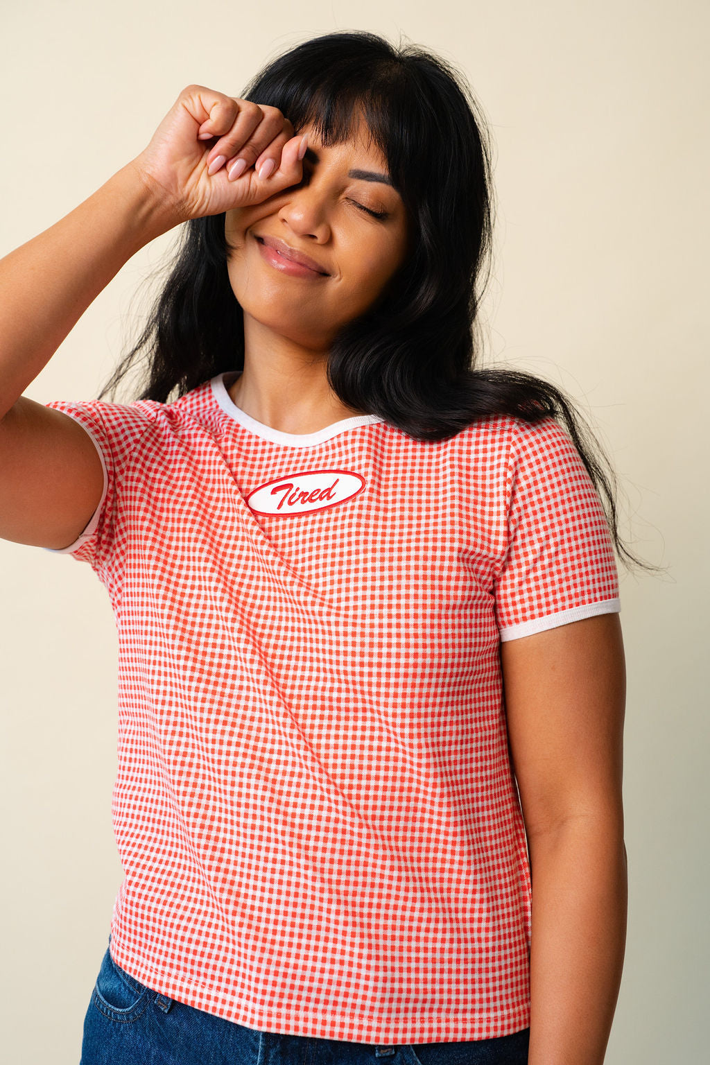 Person wearing a red and white gingham printed tee with a red and white "Tired" patch on the front, rubbing their eye.