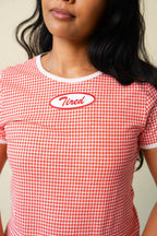 Person wearing a red and white gingham printed tee with a red and white "Tired" patch on the front. 