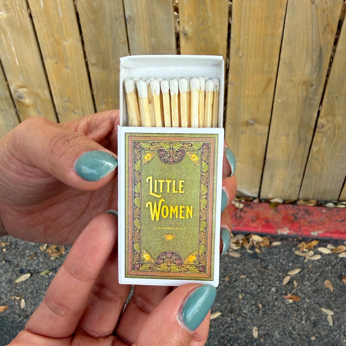 Matchbox with 'Little Women' cover held by a hand against a wooden fence background