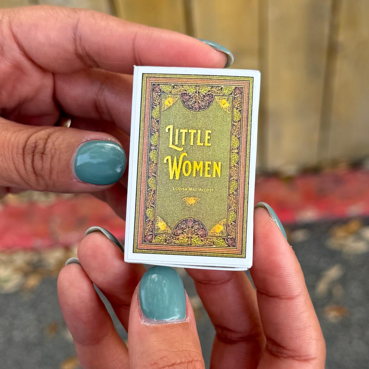 Hand holding a miniature book titled 'Little Women' with decorative cover.