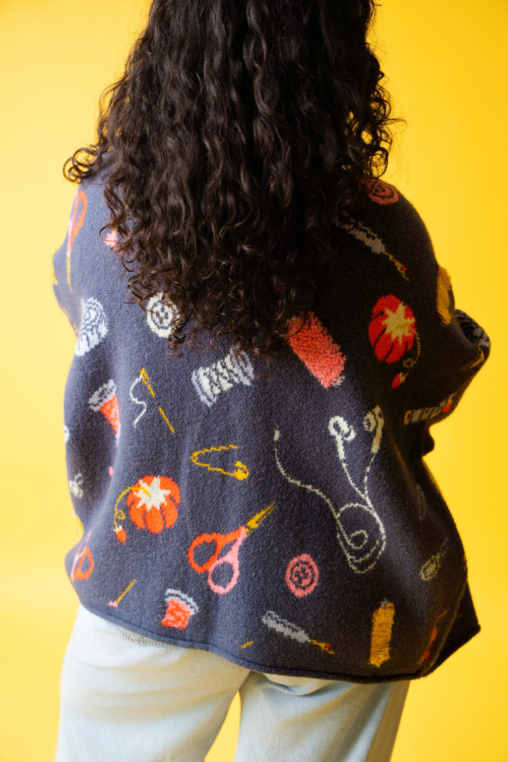 Back of person wearing a colorful sweater with sewing items on it and jeans on a yellow background. 