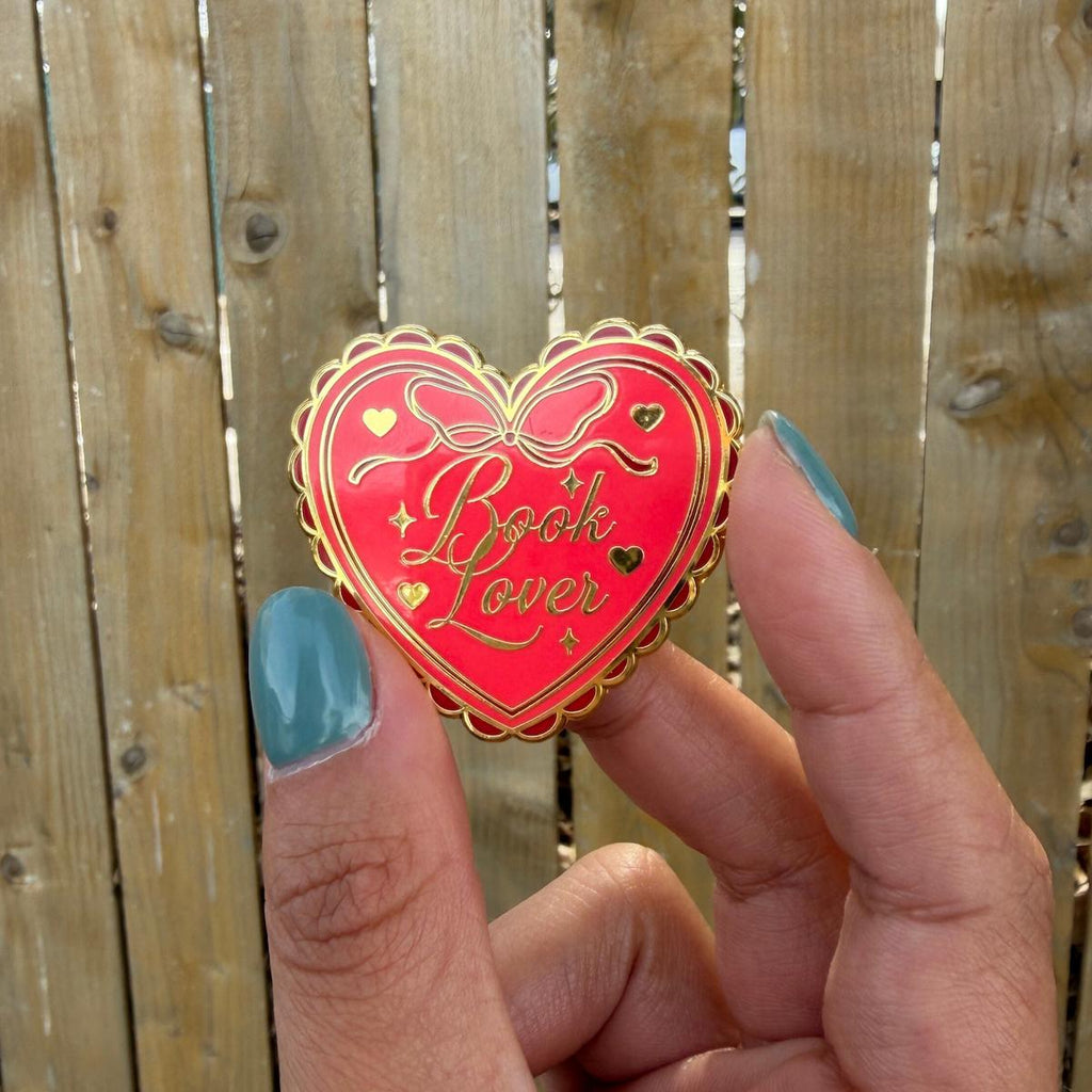 Heart-shaped 'Book Lover' pin held by a hand with blue nail polish against a wooden fence background.