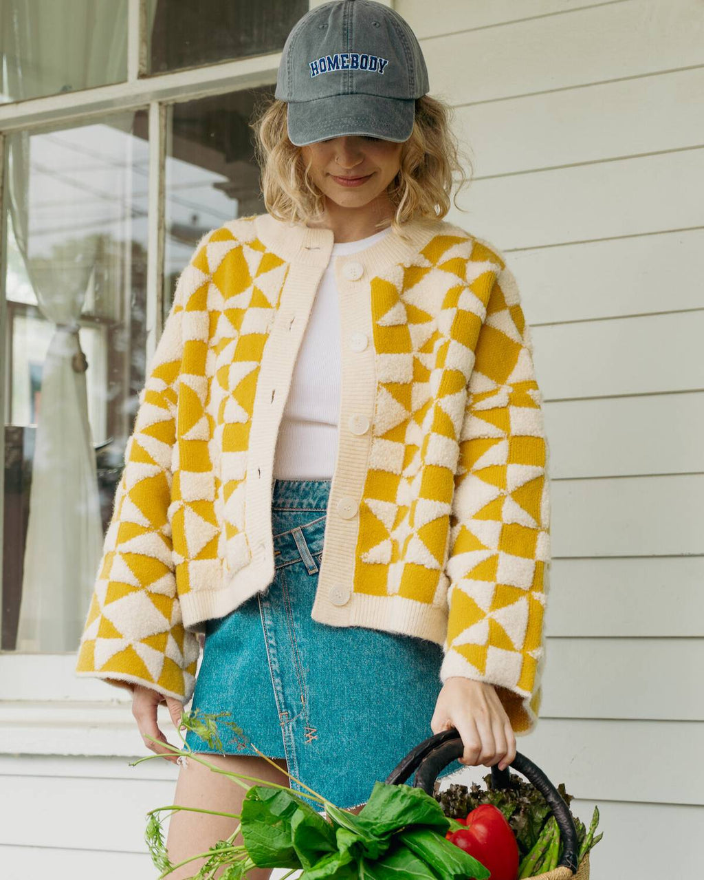 Person wearing a yellow and white patterned jacket with a blue cap, standing outside holding a basket of vegetables.