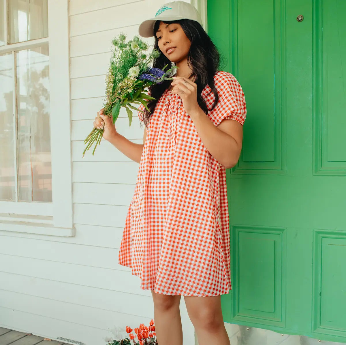 Woman in a red checkered dress holding flowers in front of a green door.