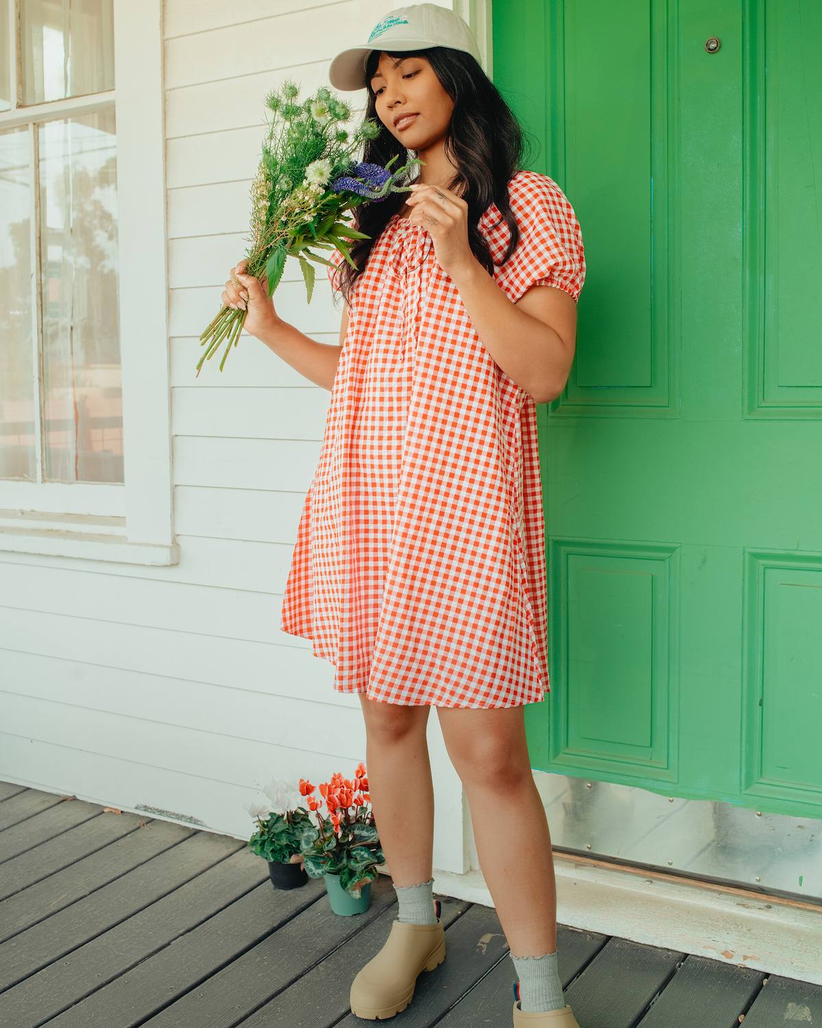 Woman in a red checkered dress holding flowers on a porch with a green door.