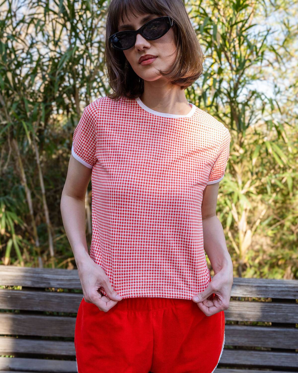 Person wearing a red checkered shirt and red pants sitting on a bench with greenery in the background