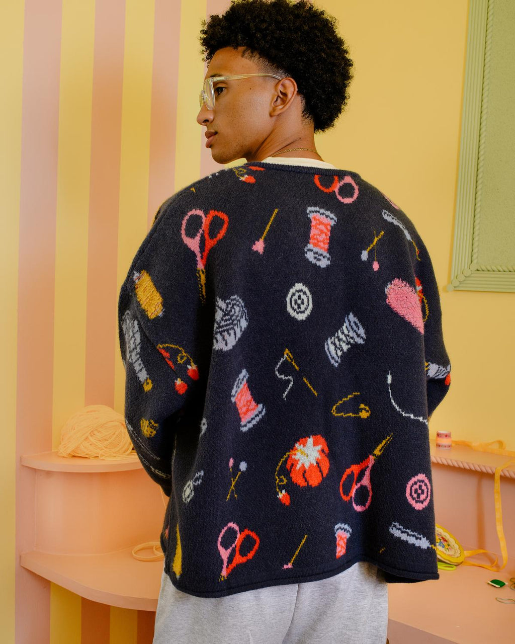 Back of person wearing a colorful sweater with sewing items on it in a pastel-colored room.