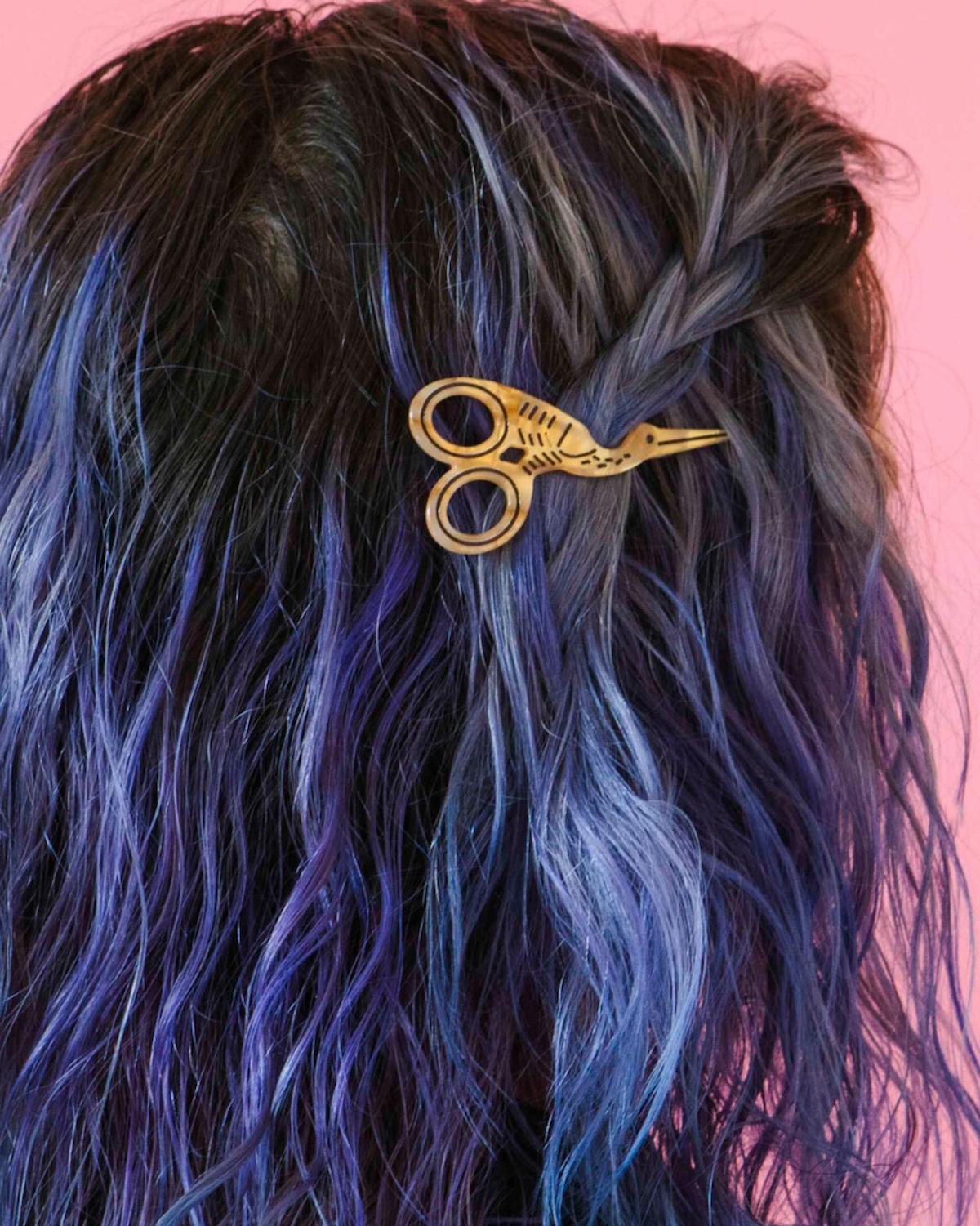 Hair accessory shaped like gold scissors on blue hair against a pink background