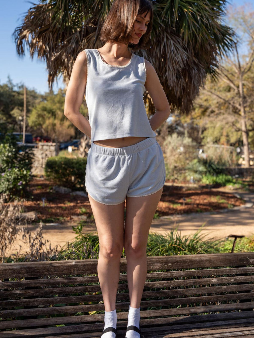 Woman in light blue sleeveless top and shorts standing on a wooden bench in a garden.