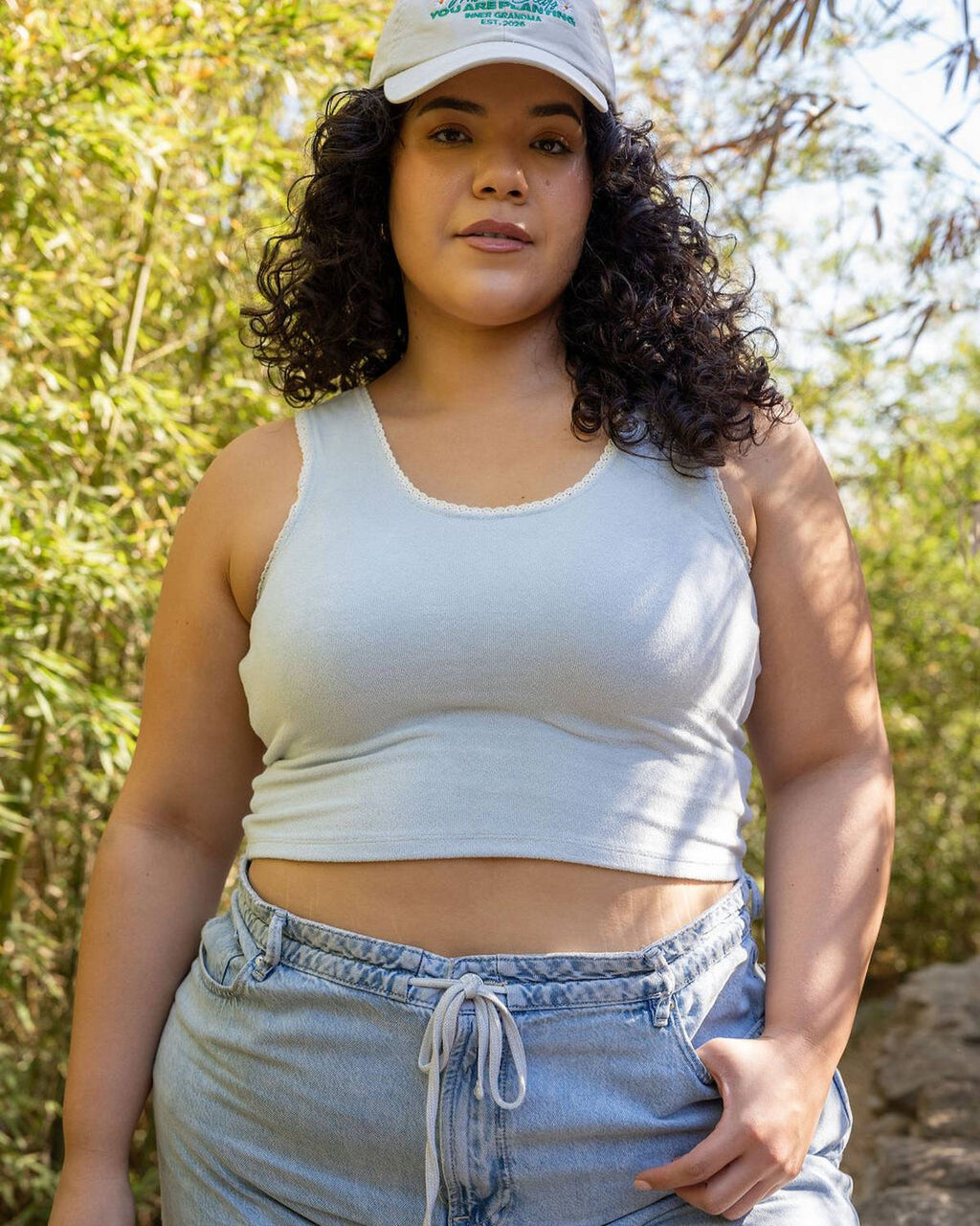 Woman wearing a white tank top and cap outdoors with greenery in the background