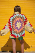 A person standing with their back to the camera, wearing a colorful quilted jacket with a large geometric starburst pattern on the back, posed in front of a bright yellow brick wall.