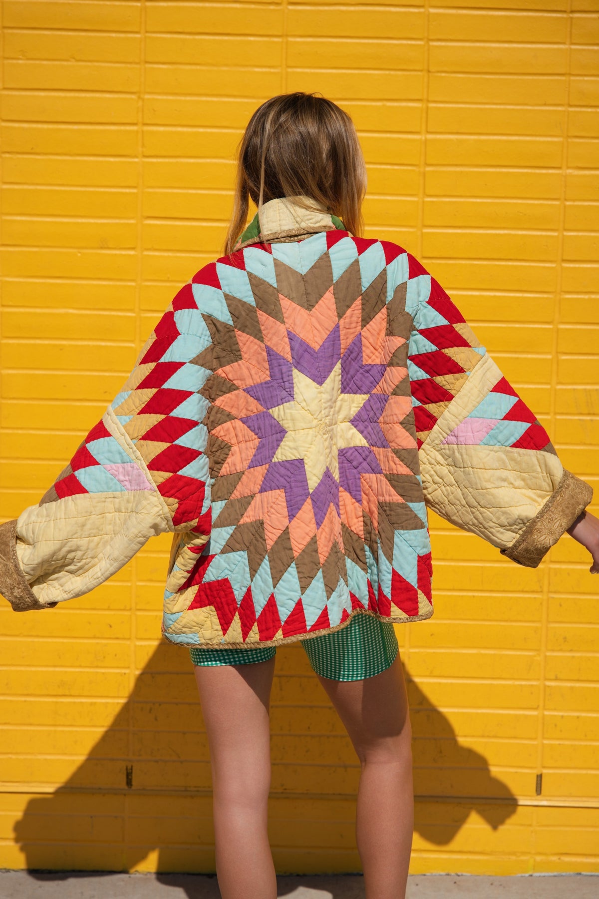 A person standing with their back to the camera, wearing a colorful quilted jacket with a large geometric starburst pattern on the back, posed in front of a bright yellow brick wall.
