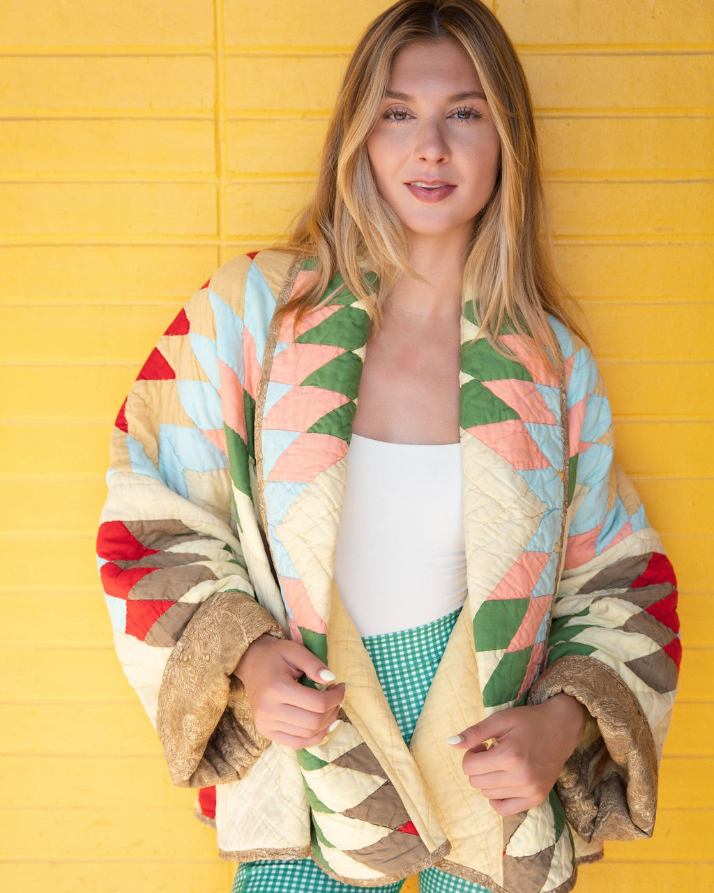 Person standing against a bright yellow paneled wall wearing a colorful geometric quilted cardigan with faux fur trim, a white tank top, and green gingham shorts.