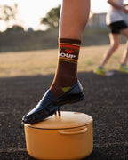 A person outdoors at sunset wearing a brown crew sock with red and yellow stripes and the text 'I heart SOUP' in white, paired with a black loafer, stepping onto a yellow pot.