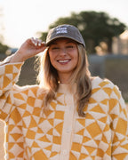 A woman standing outdoors in a field at sunset wearing a yellow and cream geometric patterned cardigan, red shorts, and a dark SOUP MODE baseball cap.