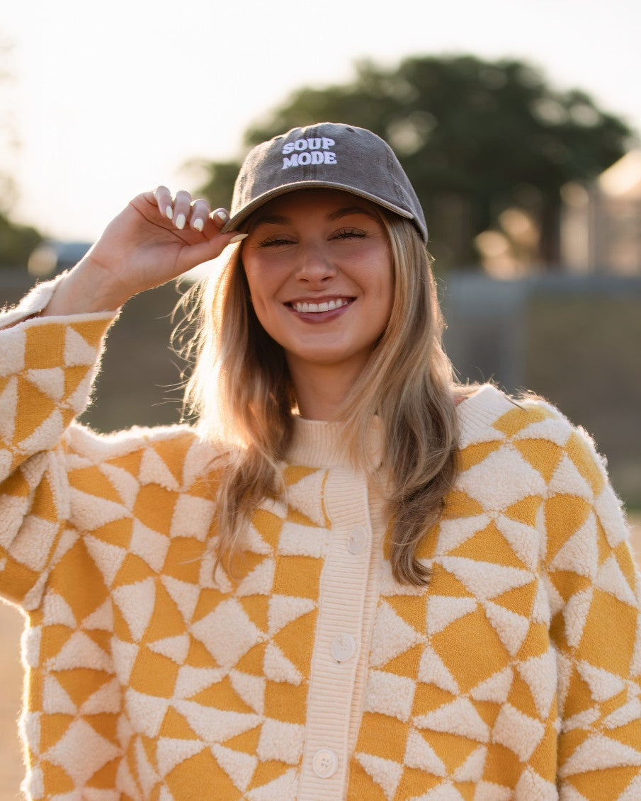 A woman standing outdoors in a field at sunset wearing a yellow and cream geometric patterned cardigan, red shorts, and a dark SOUP MODE baseball cap.