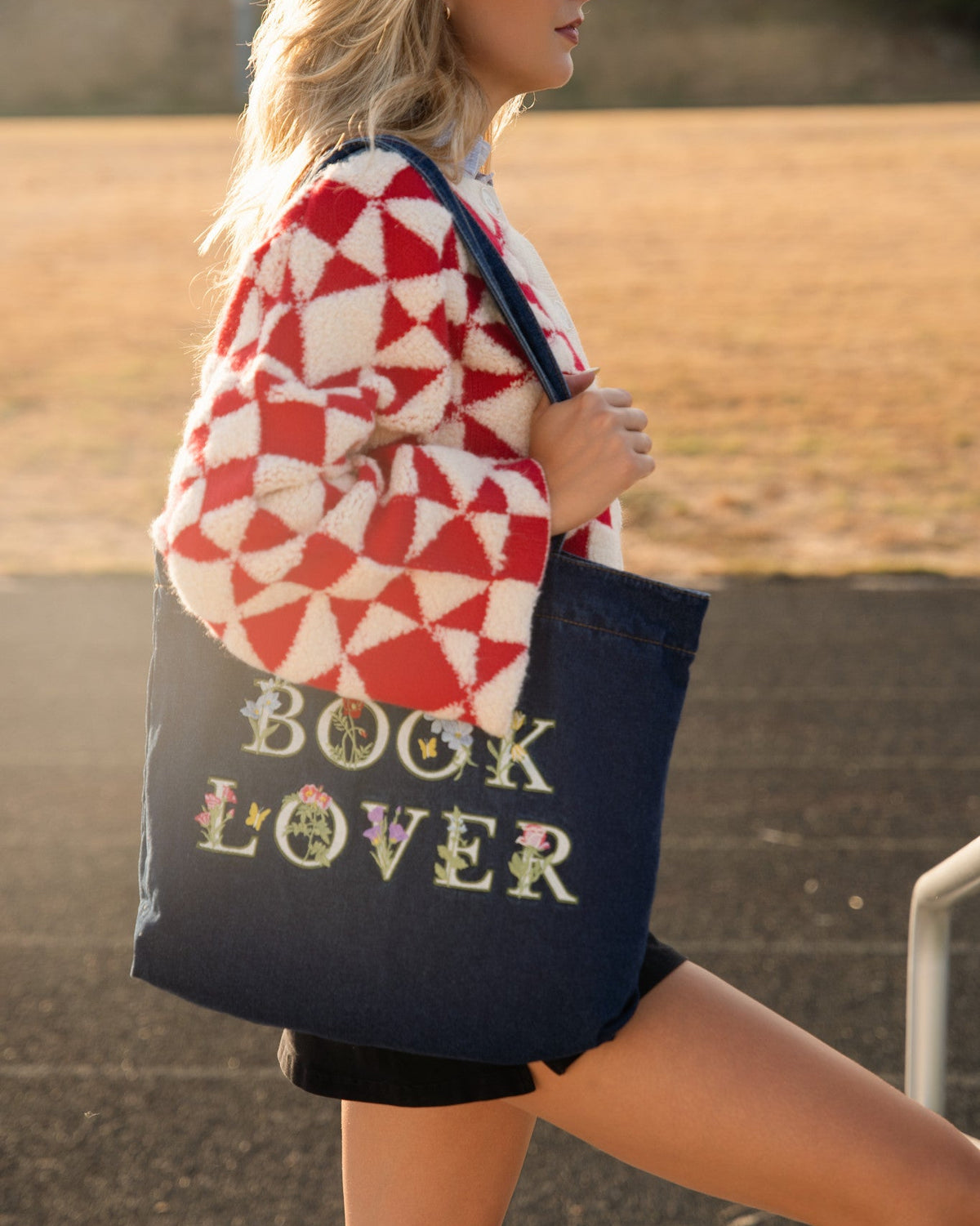 Person in a red and white patterned sweater carries a navy blue denim canvas tote bag with 'BOOK LOVER' written on it in a floral font, set against an outdoor background.