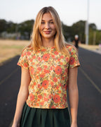 A woman standing on an outdoor track wearing a short-sleeve crewneck t-shirt with a vibrant orange, red, and yellow floral print, paired with a dark green pleated mini skirt.