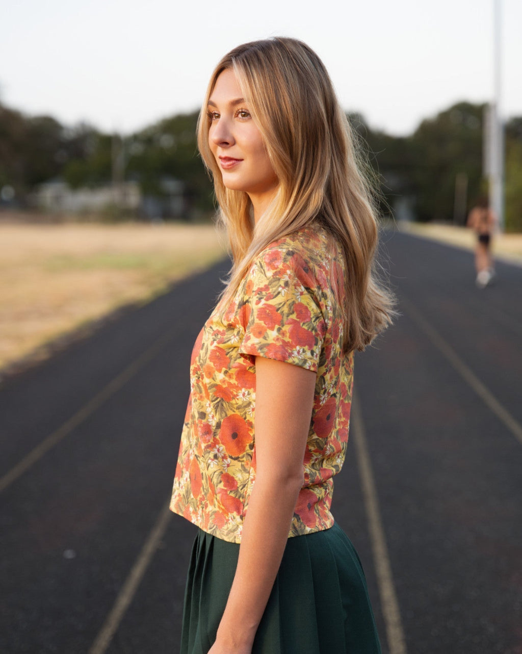 A woman standing on an outdoor running track wearing a short-sleeve orange and yellow floral shirt and a dark green pleated skirt.