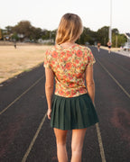 A woman standing on an outdoor running track, viewed from behind. She is wearing a short-sleeve floral top with orange, red, and yellow flowers on a beige background, paired with a dark green pleated mini skirt.