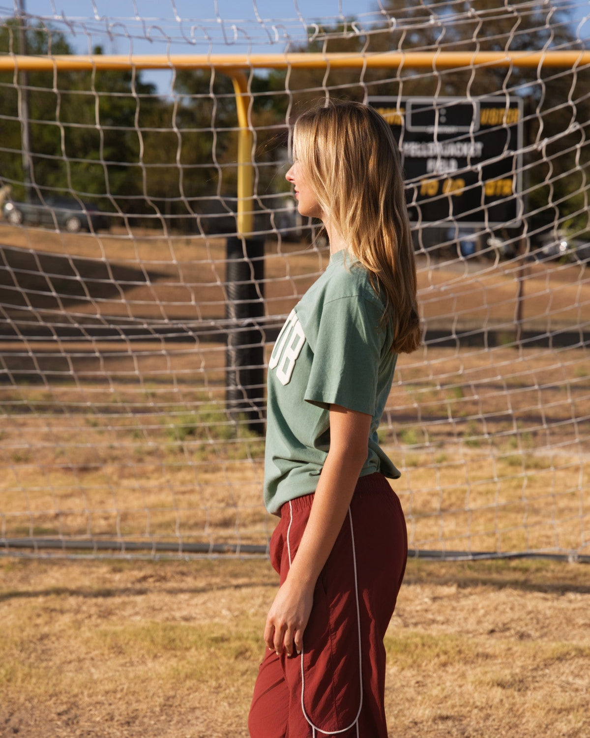 A young woman standing in profile outdoors in front of a soccer goal net, wearing a sage green t-shirt and dark red cargo pants.