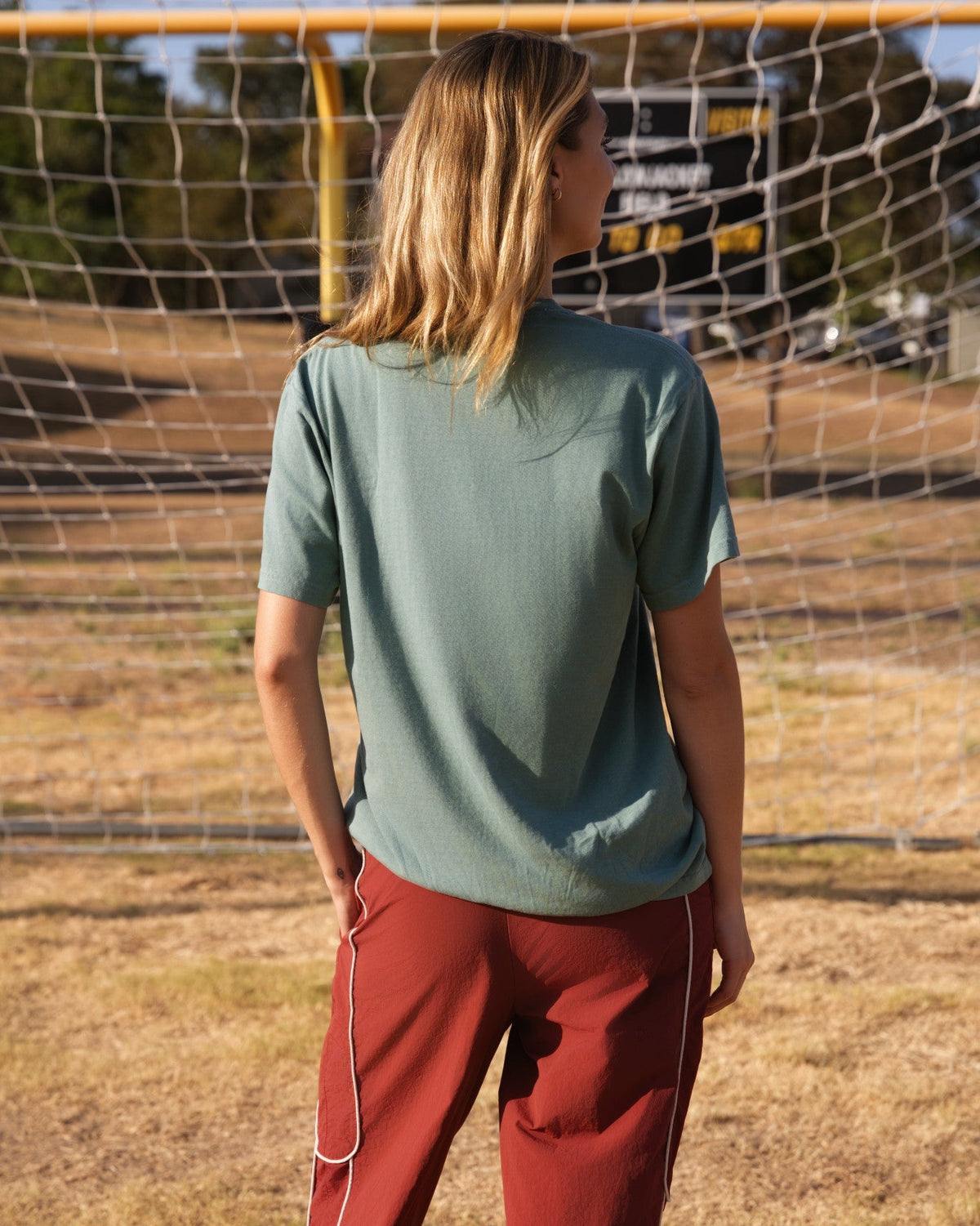 A woman standing outdoors on a dry grass field in front of a soccer goal, wearing a muted sage green short-sleeve t-shirt and rust red wide-leg pants with white piping.