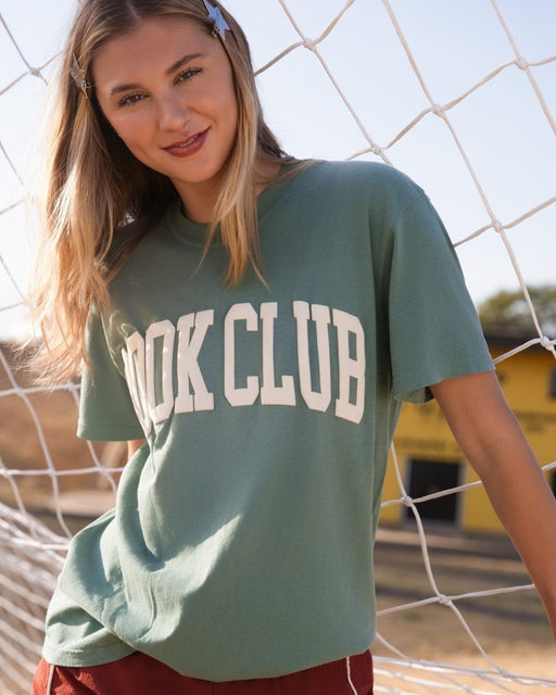 A woman wearing a sage green t-shirt with 'BOOK CLUB' printed in white varsity letters, posing in front of a white soccer net with a sunny outdoor background.