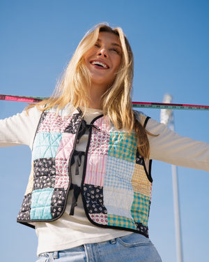 Person outdoors wearing a colorful patchwork quilted vest over a white long-sleeve top and light blue jeans, with her arms outstretched against a clear blue sky.