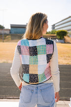 Person viewed from behind wearing a colorful patchwork quilted vest over a white long-sleeve top and light blue jeans, standing outdoors.