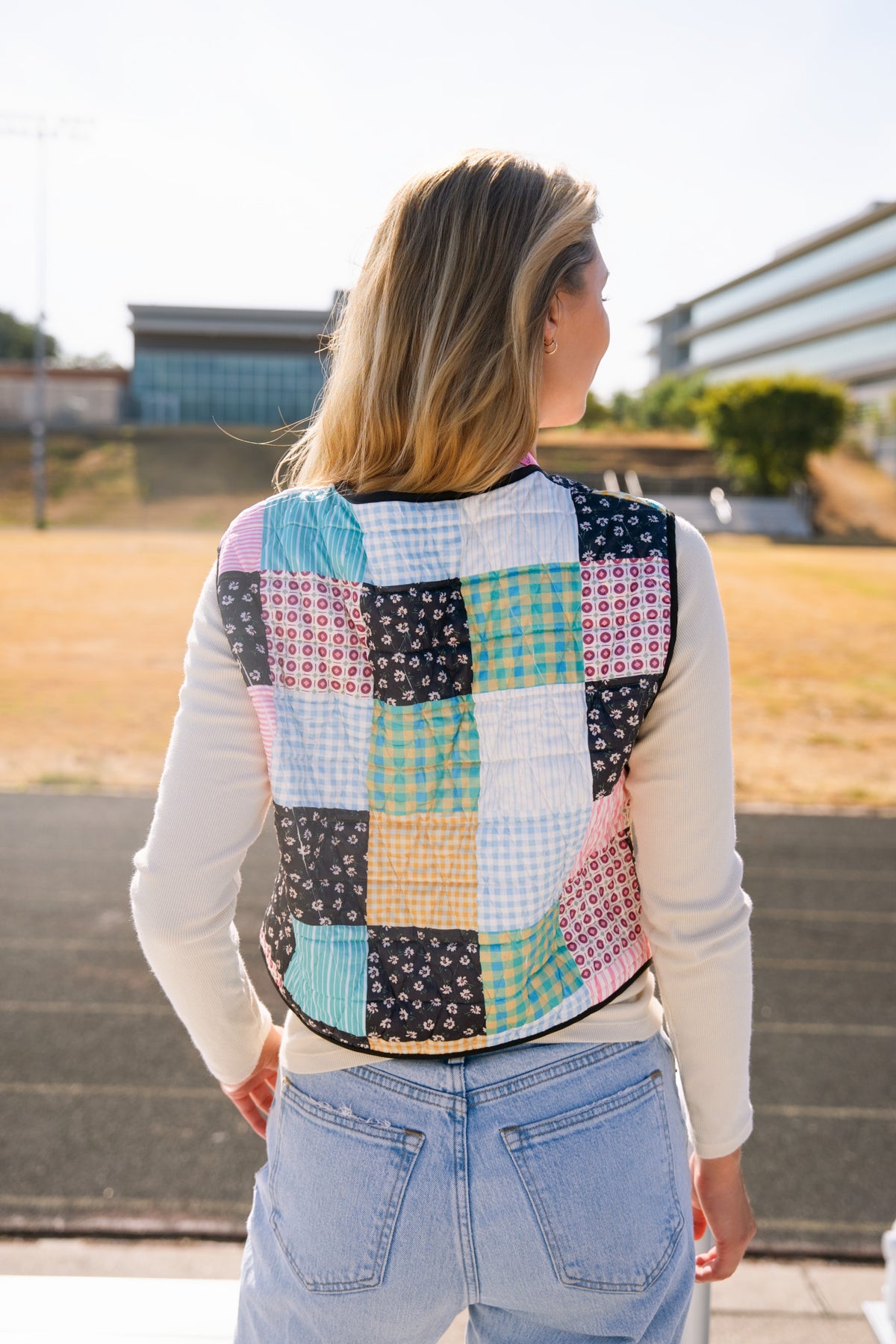 Person viewed from behind wearing a colorful patchwork quilted vest over a white long-sleeve top and light blue jeans, standing outdoors.