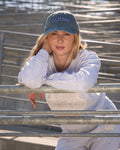 Person wearing a light gray sweatshirt and matching sweatpants with a blue baseball cap that says 'HOMEBODY' in white lettering, leaning on a metal railing in an outdoor setting.