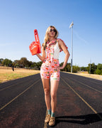 Person standing on a running track wearing a white tank top and matching floral shorts, holding a red foam finger. 