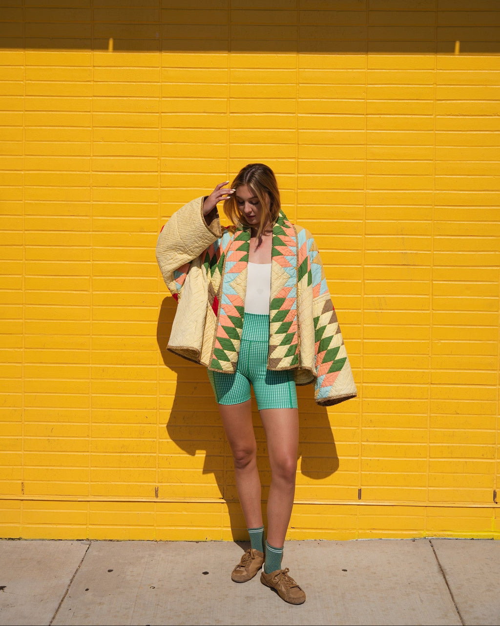 Person standing in front of a bright yellow brick wall wearing a cream knit cardigan with a colorful geometric pattern, mint green shorts, a white tank top, and brown shoes.