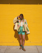 Person standing in front of a bright yellow brick wall wearing a cream knit cardigan with a colorful geometric pattern, mint green shorts, a white tank top, and brown shoes.